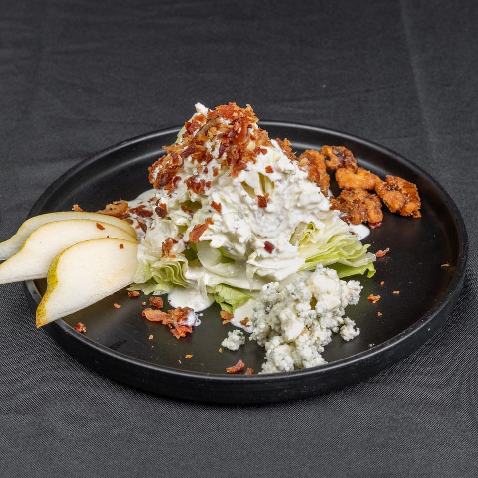 Wedge salad topped with creamy dressing, bacon, blue cheese, and lettuce, served with pear slices and fried chicken on a black plate.