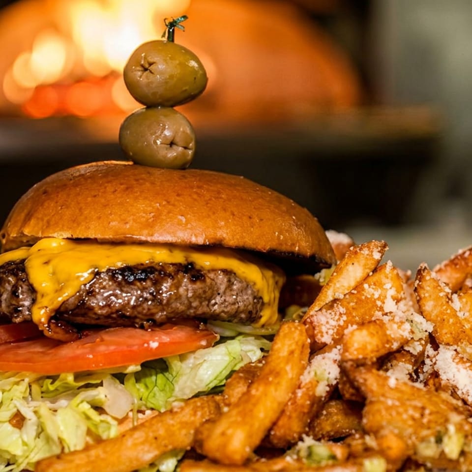 Cheeseburger topped with two olives on a skewer, served with lettuce, tomato, and seasoned fries on a plate.