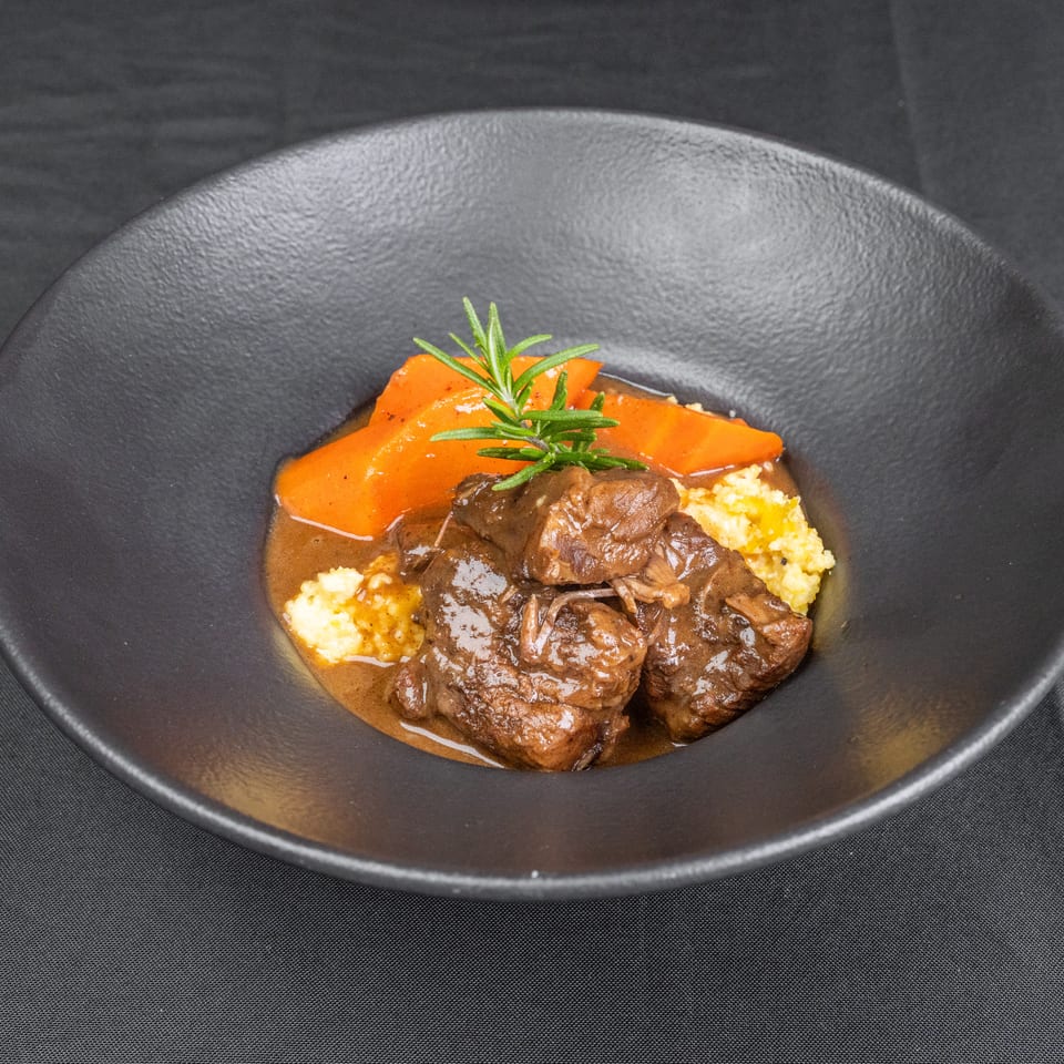 Braised beef in brown sauce served with creamy polenta and roasted carrots, garnished with fresh rosemary on a black plate.