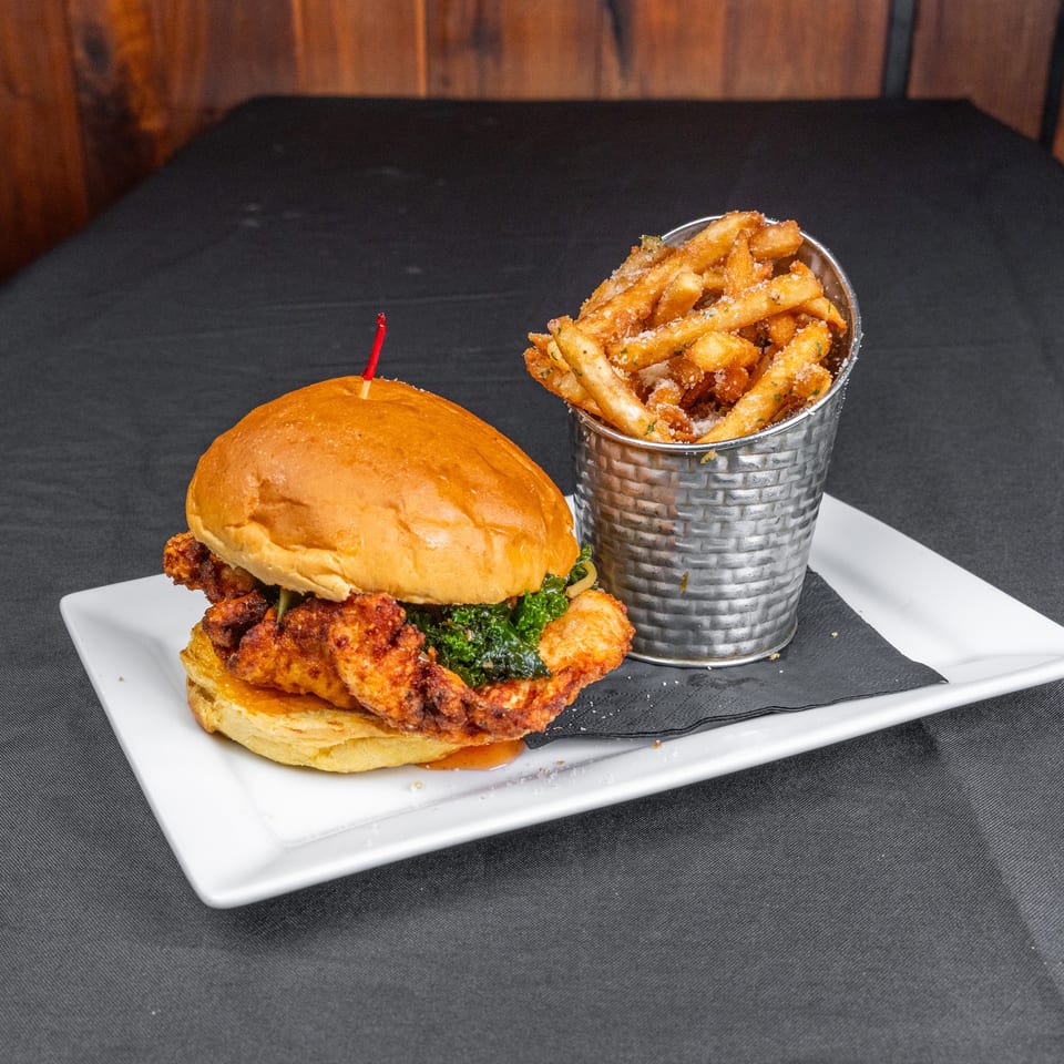 Crispy fried chicken sandwich with greens on a brioche bun, served with seasoned fries in a metal cup on a white plate.