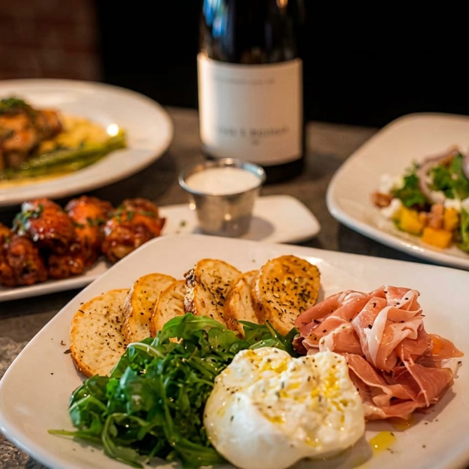 Assorted restaurant dishes include burrata with prosciutto and greens, glazed wings, salad, and a plated entrée beside wine.