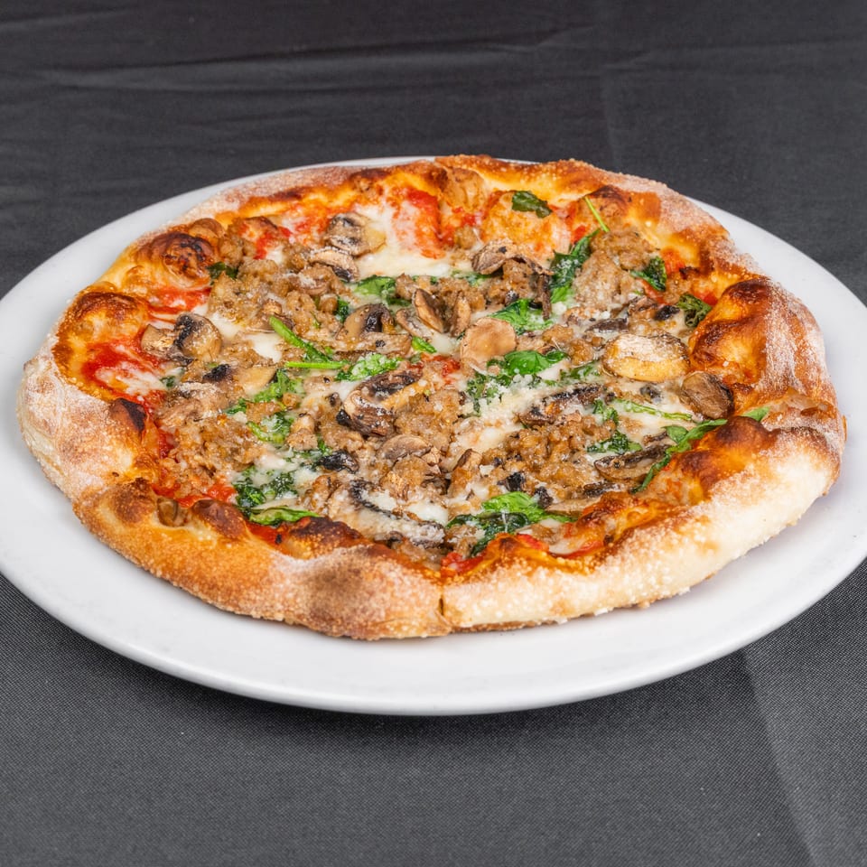 Wood-fired pizza topped with melted cheese, mushrooms, sausage, spinach, and tomato sauce on a white plate.