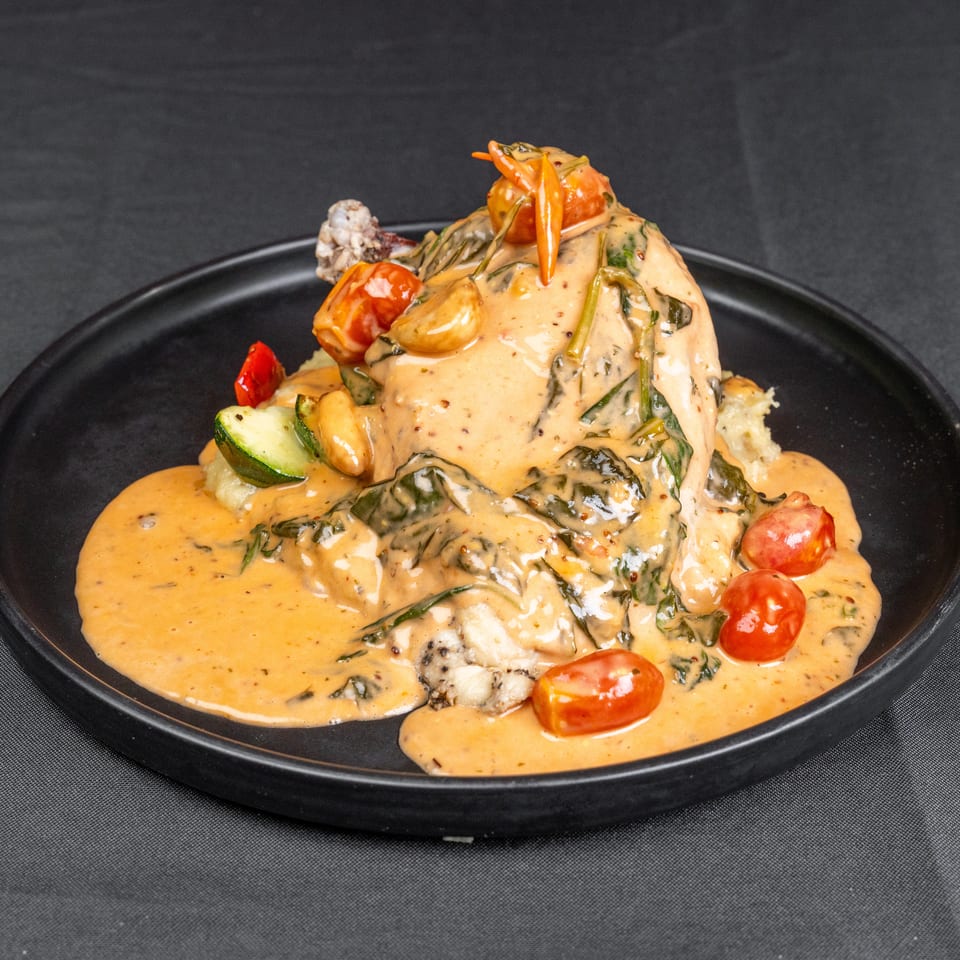 Creamy sauced entrée with vegetables, cherry tomatoes, herbs, and mashed potatoes served on a black plate.