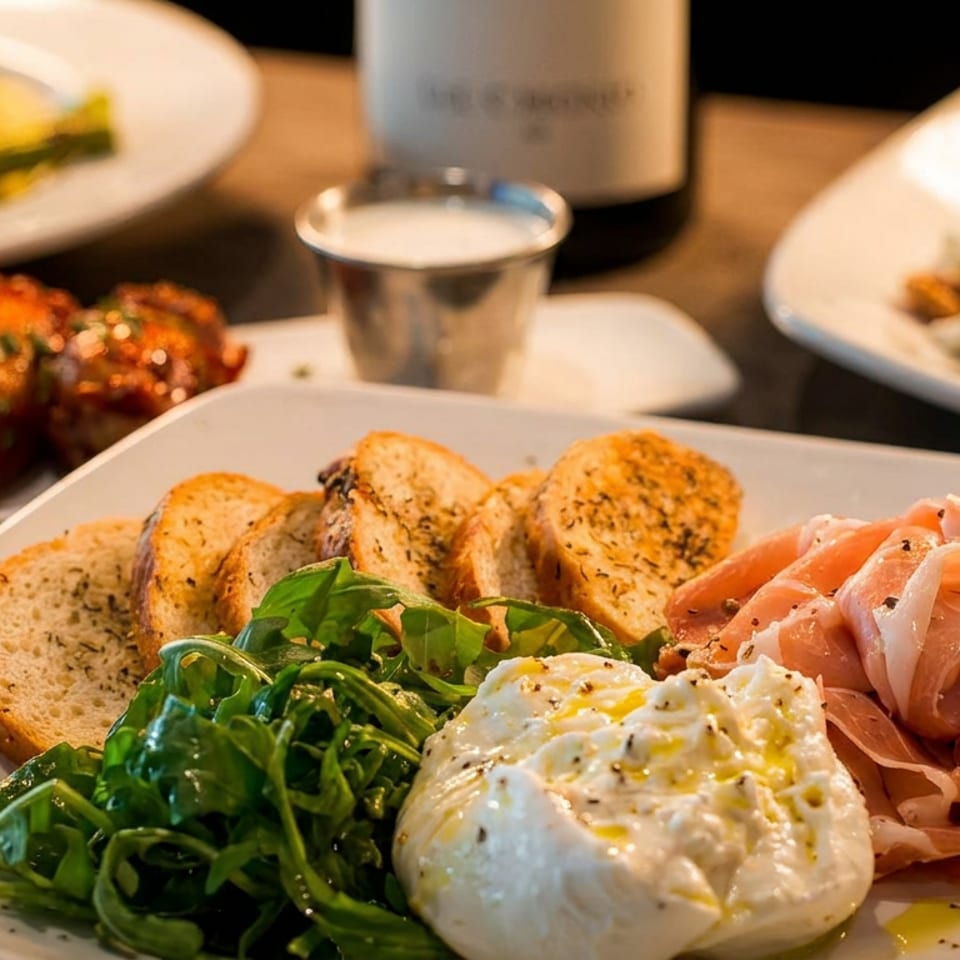 Italian appetizer platter with burrata, prosciutto, arugula, toasted bread, and roasted tomatoes on a restaurant table.