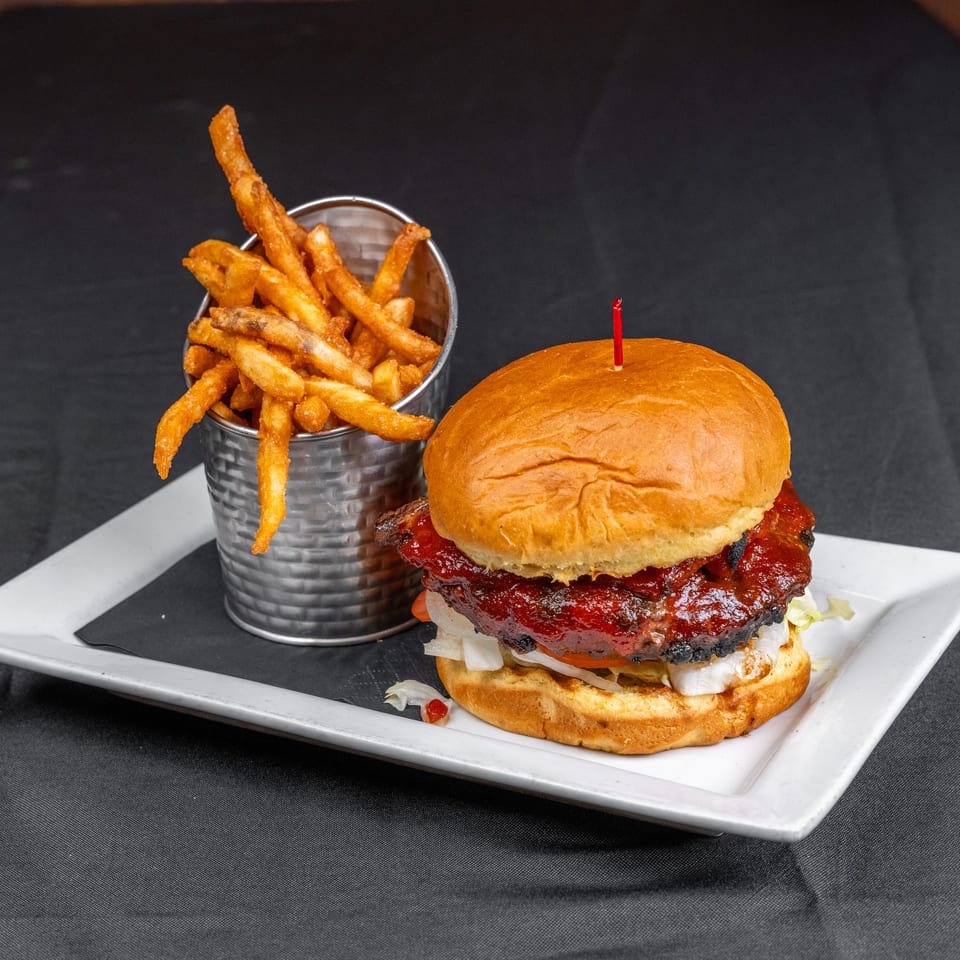 Barbecue burger with lettuce and tomato served alongside a metal cup of golden fries on a white plate.